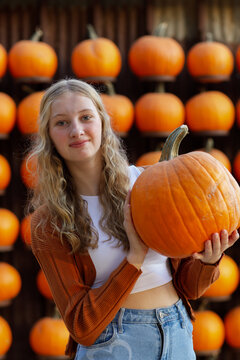 portrait of a blonde teenage girl happily holding a pumpkin in front of a wall of pumpkins