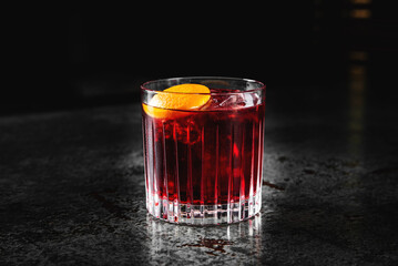 A vibrant red cocktail in a short glass, garnished with an orange slice, set against a dark...