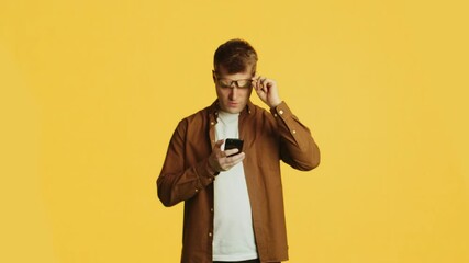 Confused man putting on glasses reading bad news on smartphone on yellow background