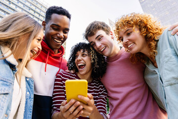Happy young group of diverse teenage friends using mobile phone at city street. Generation Z people having fun watching media content on social platform app outside. Youth community concept