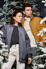 A joyful couple stands together in winter jackets, surrounded by enchanting pine trees dusted in...