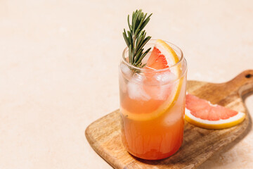 Grapefruit cocktail with rosemary garnish in a glass, served on a wooden cutting board. Vibrant, refreshing drink perfect for summer menu or cocktail recipes. Greyhound, lemonade, paloma, mocktail.