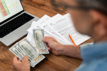 Naklejka premium Man reviewing tax documents while holding dollar bills near a laptop on a wooden desk. Financial management, tax refund, and accounting concept. 