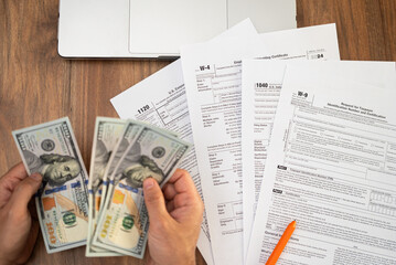 Person counting hundred-dollar bills while reviewing tax forms on a wooden desk with a laptop nearby. Tax preparation and financial planning concept, ideal for accounting and finance themes 