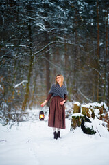 Naklejka premium Portrait of beautiful blonde girl in medieval dress and woolen scarf in winter snowy forest. Young worker in rural scene with flashlight.Colorful fairytale art work.