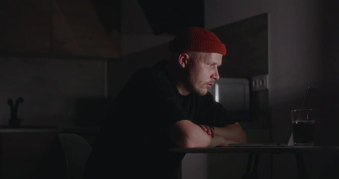 A millennial man in a dark T-shirt works from his kitchen using a laptop. The millennial introvert works at night from his home. A difficult project at work that requires round-the-clock work. 4k