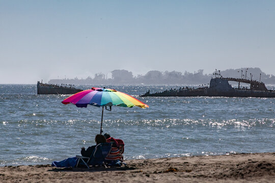 Couple reclining under Rainbow colored beach umbrella at Seacliff State Beach,  with concrete ship (SS Palo Alto) in background on a beautiful, sunny day in Aptos, California
