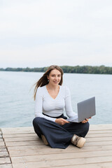 A young woman with long brown hair is sitting on a wooden pier by the lake, working on a laptop. She is dressed in a white, long-sleeved top and wide-leg navy pants, looking relaxed and approachable. 