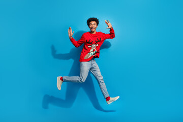 Full body photo of attractive young man running jump excited dressed red christmas sweater theme party on blue color background