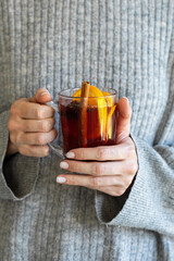 A cozy close-up of hands holding a glass mug of warm mulled wine with cinnamon and orange slices.