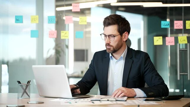 Busy financier in formal suit is doing paperwork using a laptop computer while sitting at workplace. Confident businessman working fills out documents, writing, making a tax return in business office