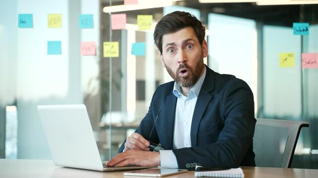Happy businessman in a formal suit received great news on a laptop computer, surprised looking camera sitting at the desk at workplace in business office. Man shocked, rejoices. Wow, amazed, success