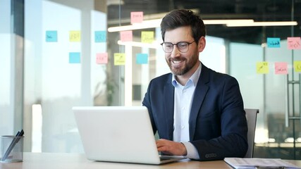Businessman working at desk in modern office using laptop. Smiling man focused, planning business strategy in contemporary workplace. Manager in glasses working in computer application, banking