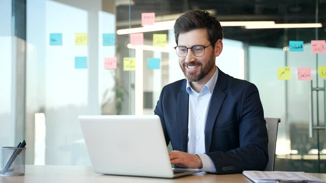 Businessman working at desk in modern office using laptop. Smiling man focused, planning business strategy in contemporary workplace. Manager in glasses working in computer application, banking