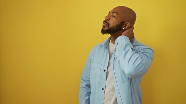 Handsome young man with a beard and bald head wearing a light blue shirt touches his neck in discomfort against an isolated yellow background