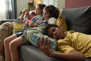 Group of teenagers lounging on sofa, immersed in using their smartphones. Cozy living room setting with relaxed atmosphere and modern furniture