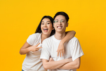 Fototapeta premium Portrait of joyful asian couple embracing over yellow studio background, girl showing peace symbol
