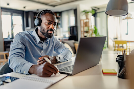 African-American man wearing headphones, taking notes while working on a laptop from home, focused and engaged in a modern workspace.