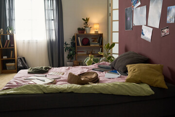 Obraz premium Cozy bedroom interior featuring bed with pillows and blanket, small side table with lamp, books, and potted plants on shelves, photographs on the wall, natural light streaming in