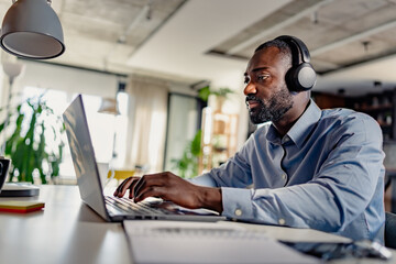 Obraz premium Focused man wearing headphones, working on his laptop in a modern, well-lit home office environment.