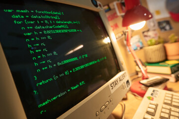 Lines of code displayed on retro computer screen with blurred background of workspace featuring books, lamp, and other desk items suggesting software development