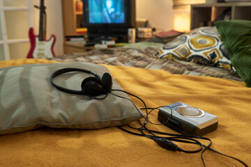 Headset and retro cassette player lying on bed in cozy room with blurred background showing guitar, television, bookshelf, and decorative pillow