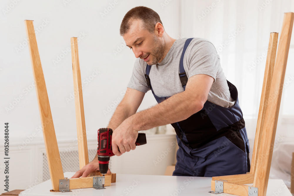 Poster carpentry service. repairman in blue uniform fixing table with electric drill standing indoors. furn