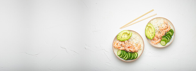 Two white ceramic bowls with rice, shrimps, avocado, vegetables and sesame seeds and wooden sticks on white concrete stone background top view. Healthy asian style poke bowl, space for text.