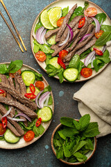 Two plates with traditional Thai beef salad with vegetables and mint top view served on rustic concrete background, healthy exotic asian meal.