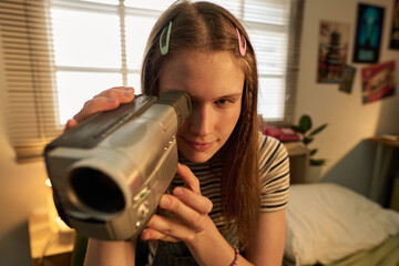 Young woman engaged in filming with retro camcorder, seated on bed in cozy bedroom setting with...