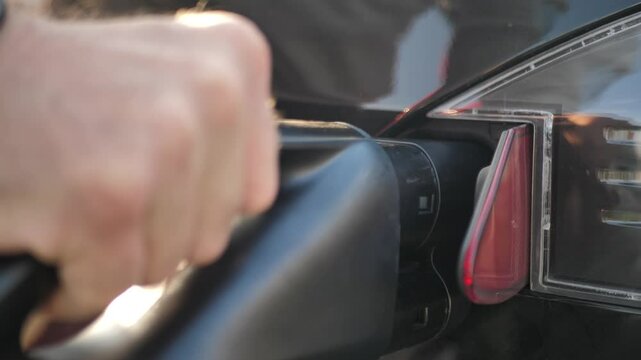 A man's hand holding a gun with a plug of an electric car charger inserts it into a socket to recharge a hybrid car. Using environmentally friendly electricity and alternative sources