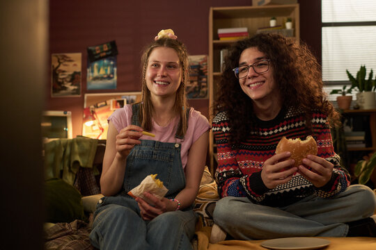 Two friends are sharing burgers in cozy retro living room setting, enjoying relaxed atmosphere with warm lighting and casual attire, smiling and having a good time