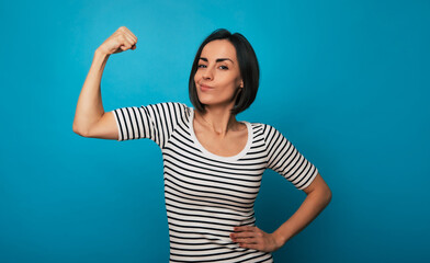 A young woman in a striped shirt flexes her arm with a confident expression against a blue...
