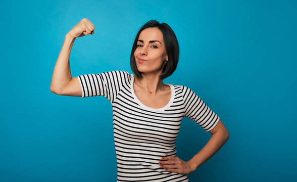 A young woman in a striped shirt flexes her arm with a confident expression against a blue background. Her pose conveys strength, empowerment, and self-confidence, embodying a "can-do" attitude.
