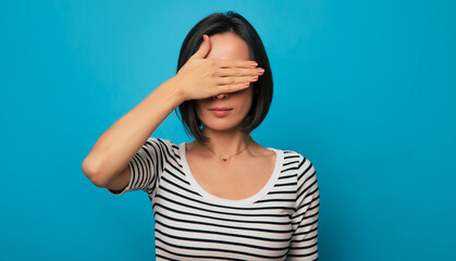 A young woman in a striped shirt covers her eyes with one hand against a blue background. Her gesture suggests mystery, avoidance, or playfulness, adding an element of curiosity and intrigue.