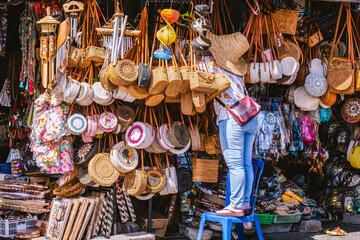 Tourist standing on a blue plastic chair, choosing a handmade rattan bag in a colorful souvenir shop at the Tanah lot temple market