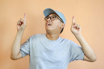 Fototapeta premium Excited and fascinated asian delivery guy in grey cap and t-shirt smiling amazed, looking and pointing fingers up, showing top advertisement with super cool discounts, white background