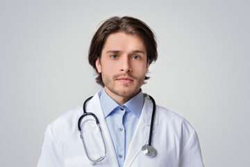 Fototapeta premium Portrait Of Handsome Young Doctor Wearinf White Medical Coat And Stethoscope And Looking To Camera, Light Studio Background, Copy Space