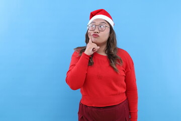 Merry young Asian woman wear red turtleneck hat posing put hand prop up on chin, lost in thought and conjectures isolated on blue background. Happy New Year 2025 celebration Christmas holiday concept