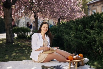 young woman, casual picnic, picnic snacks. Young woman relaxes on picnic blanket beneath cherry...