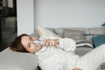 Young Woman Using Nebulizer Inhaler Lying On Sofa At Home, Suffering From Respiratory Illness Or...