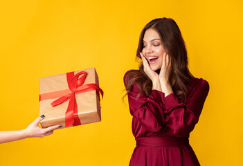 Surprise For You. Excited young lady in evening dress getting unexpected gift box, touching cheeks in joy, standing over colorful background, free space