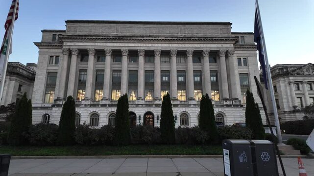 A white marble building with columns, windows, and a sign reading United States Department of Agriculture. There are bushes and trees in front of the building. The sky is blue and there is a flagpole 