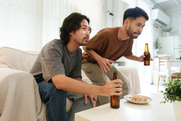 Two Asian men drinking beer in a living room while watching football games on television intently with focused expressions. Two friends with soccer game. Friendship concept