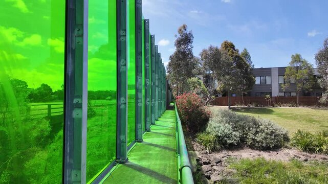 A modern green glass sound barrier wall or transparent noise protection panels line the busy highway, creating a landscaped border that separates the residential neighborhood houses and traffic.