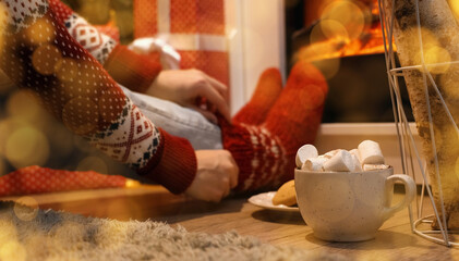 Woman with warm socks and cup of hot chocolate sitting near fireplace in festive living room at evening