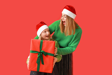 Cute little girl and her mother in Santa hats with Christmas gift box on red background