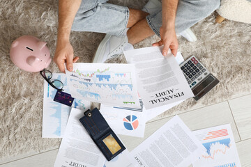 Young man in debt with documents sitting on floor at home, top view