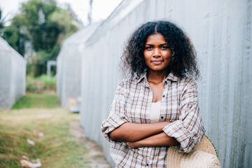 Portrait of a confident woman farmer in a checkered shirt and apron arms crossed holding a hat smiling in front of a farm's lush greenhouse tomatoes. Nature's beauty in a farming setting.