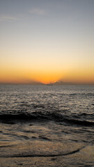 A calm seascape displays the bright colors of a tropical sunset, with waves gently lapping the shore and distant clouds illuminated by the last rays of the sun, creating a peaceful beach scene.